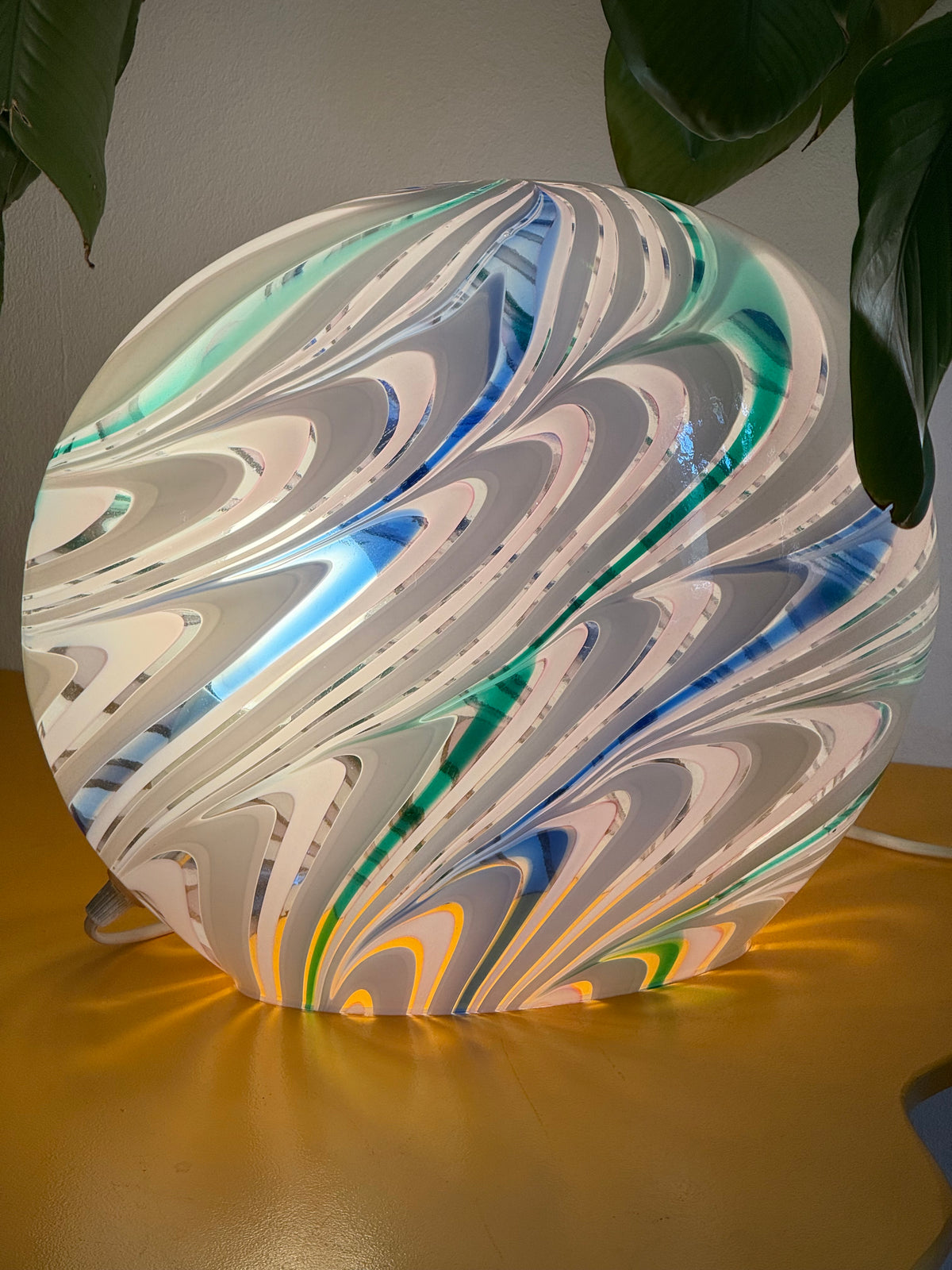 Tændt rund Murano bordlampe i glas med blå og grøn swirl foran grønne planter